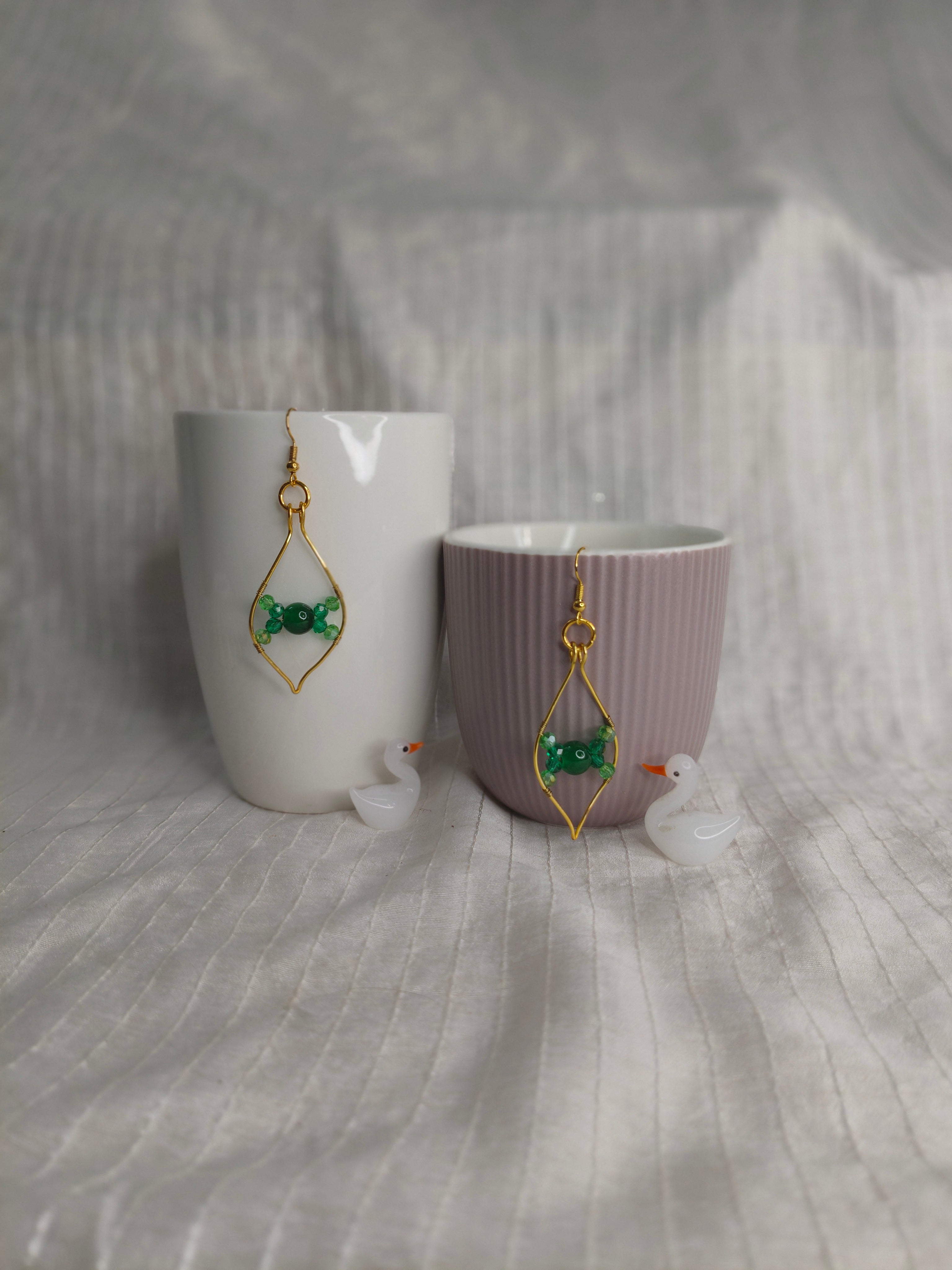 Pair of earrings with green gemstones and gold accents displayed on a white and purple cup against a textured gray background.