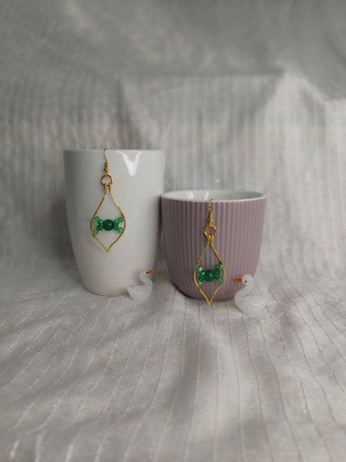 Pair of earrings with green gemstones and gold accents displayed on a white and purple cup against a textured gray background.